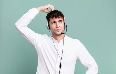 Yetişkin bir adam kafası karışmış ve kafası karışmış hissediyor, kulaklıkla kafasını kaşıyor. tele pazarlamacı konsepti
