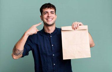 Kendine güvenen yetişkin bir adam kahvaltı poşeti ve paket kağıdıyla kendi geniş gülüşünü işaret ediyor.