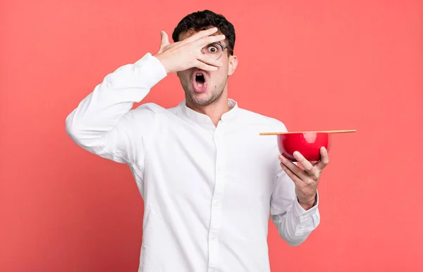 adult man looking shocked, scared or terrified, covering face with hand holding a ramen noodles bowl
