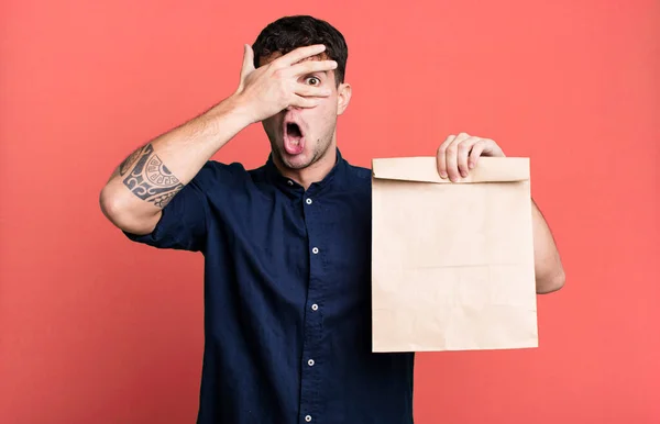 adult man looking shocked, scared or terrified, covering face with hand with a take away breakfast paper bag with a take away breakfast paper bag