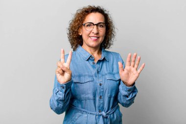 Orta yaşlı İspanyol bir kadın gülümsüyor ve dost canlısı görünüyor. Elleri önde, geriye doğru sayıyor.