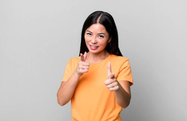 Güzel Latin bir kadın, pozitif, başarılı, mutlu bir tavırla gülümsüyor kamerayı işaret ediyor, elleriyle silah işareti yapıyor.