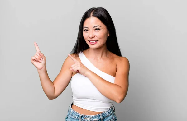 pretty latin woman smiling happily and pointing to side and upwards with both hands showing object in copy space