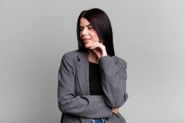 Genç ve güzel bir kadın mutlu ve kendinden emin bir ifadeyle gülümsüyor. Elleri çenesinde, merak ediyor ve yan tarafa bakıyor.