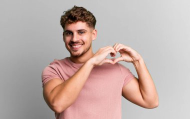 young adult caucasian man smiling and feeling happy, cute, romantic and in love, making heart shape with both hands