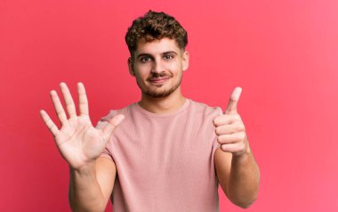 young adult caucasian man smiling and looking friendly, showing number six or sixth with hand forward, counting down