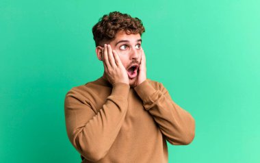 young adult caucasian man feeling happy, excited and surprised, looking to the side with both hands on face