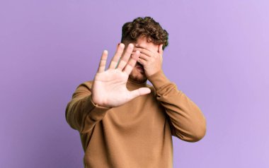 young adult caucasian man covering face with hand and putting other hand up front to stop camera, refusing photos or pictures
