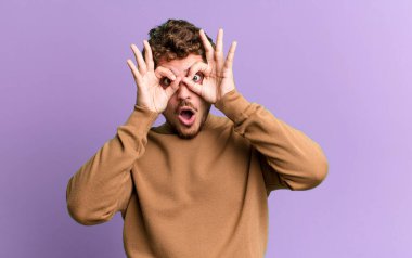 young adult caucasian man feeling shocked, amazed and surprised, holding glasses with astonished, disbelieving look