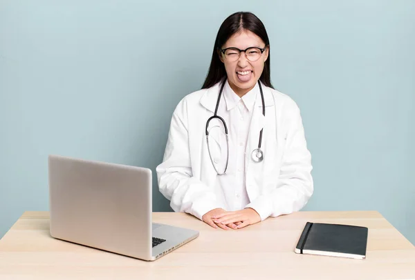 陽気で反抗的な態度の可愛いアジア系女性が冗談を言って舌を出してる 医者の机とノートパソコン — ストック写真