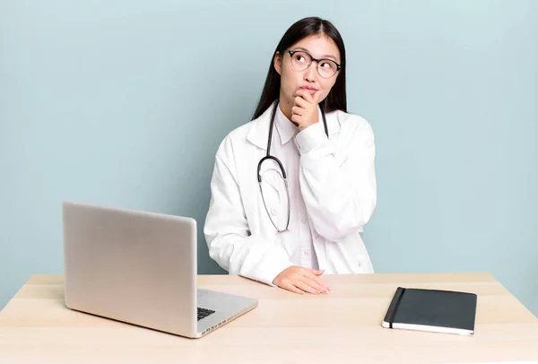 pretty asian woman thinking, feeling doubtful and confused. physician desk and laptop