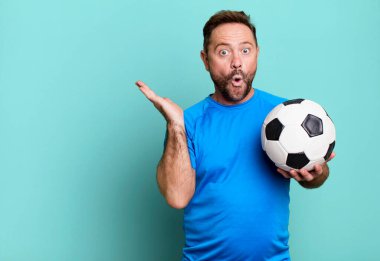 Orta yaşlı bir adam şaşırmış ve şok olmuş görünüyor. Çenesi düşmüş ve elinde bir nesne var. Bir futbol topuyla. fitness konsepti