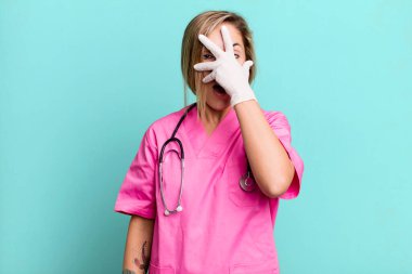 pretty blonde woman looking shocked, scared or terrified, covering face with hand. nurse concept