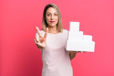 pretty blonde woman smiling and looking friendly, showing number two with a set of blank packagings