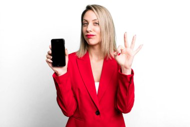 pretty blonde woman feeling happy, showing approval with okay gesture. businesswoman showing a phone screen