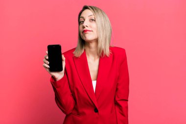 pretty blonde woman shrugging, feeling confused and uncertain. businesswoman showing a phone screen