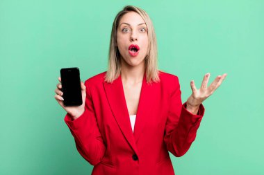pretty blonde woman feeling extremely shocked and surprised. businesswoman showing a phone screen