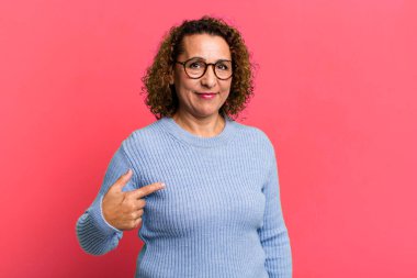 Orta yaşlı İspanyol kadın gururlu, kendinden emin ve mutlu görünüyor, gülümsüyor ve kendini işaret ediyor ya da bir numaralı işareti yapıyor.