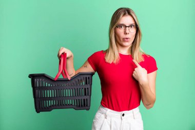 Şok olmuş ve ağzı açık, kendini gösteren genç ve güzel bir kadın. boş alışveriş sepeti kavramı