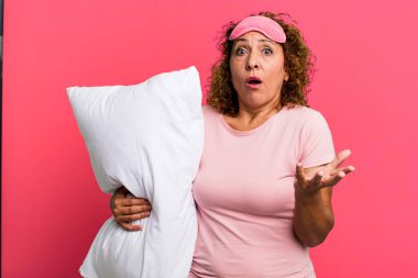 pretty middle age woman amazed, shocked and astonished with an unbelievable surprise wearing pajamas night wear and a pillow