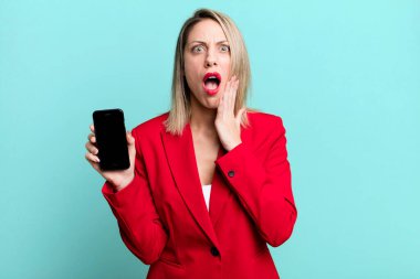 pretty blonde woman feeling shocked and scared. businesswoman showing a phone screen