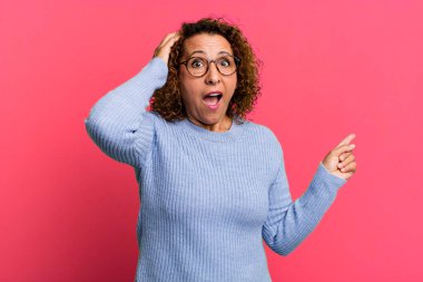 Orta yaşlı İspanyol kadın gülüyor, mutlu, pozitif ve şaşırmış görünüyor, yanal fotokopi alanını işaret eden harika bir fikrin farkına varıyor.