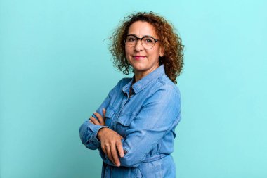 Orta yaşlı İspanyol kadın kameraya çapraz kollarıyla gülümsüyor ve mutlu, kendinden emin, tatmin olmuş bir yüz ifadesi var.