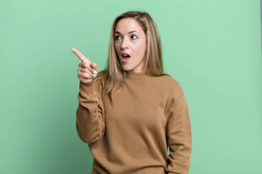 blonde adult woman feeling shocked and surprised, pointing and looking upwards in awe with amazed, open-mouthed look