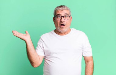 Orta yaşlı bir adam şaşırmış ve şoka uğramış görünüyor, çenesi düşmüş ve eli açık bir nesneyi tutuyor.