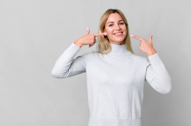 caucasian blonde woman smiling confidently pointing to own broad smile. copy space concept