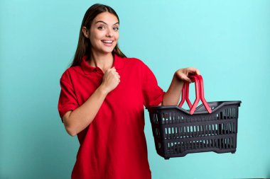 Genç ve güzel bir kadın kendini mutlu hissediyor ve bir meydan okumayla yüzleşiyor ya da kutluyor. boş alışveriş sepeti kavramı
