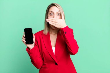 pretty blonde woman covering mouth with hands with a shocked. businesswoman showing a phone screen