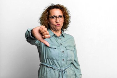 Orta yaşlı Latin bir kadın sinirli, sinirli, hayal kırıklığına uğramış ya da hoşnutsuz, ciddi bir bakışla başparmağını kaldırıyor.