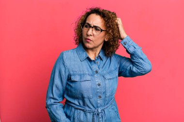 Orta yaşlı İspanyol kadın şaşkın ve şaşkın hissediyor, kafasını kaşıyor ve yan tarafa bakıyor.