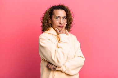 Orta yaşlı İspanyol bir kadın ciddi, düşünceli ve güvensiz görünüyor, bir kolu çapraz ve çenesi üzerinde, seçenekleri tartıyor.