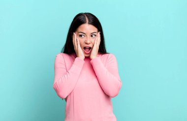 Güzel Latin bir kadın mutlu, heyecanlı ve şaşırmış hissediyor, iki eli yüzü üzerinde yana bakıyor.