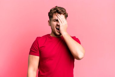 young adult caucasian man looking sleepy, bored and yawning, with a headache and one hand covering half the face