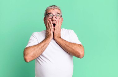 Orta yaşlı bir adam şok olmuş ve korkmuş hissediyor, ağzı açık ve elleri yanaklarında korkmuş görünüyor.