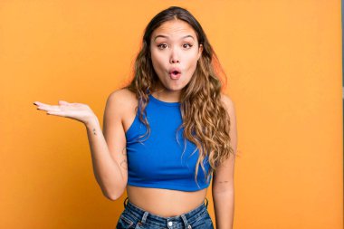 Genç ve güzel bir kadın şaşırmış ve şok olmuş görünüyor, çenesi düşmüş, bir objeyi elinde açık bir elle tutuyor.