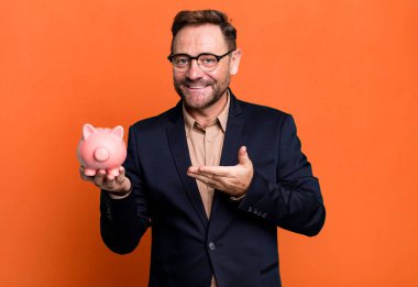 Orta yaşlı bir adam neşeli bir şekilde gülümsüyor, mutlu hissediyor ve kumbarayla bir konsept gösteriyor. iş kavramı