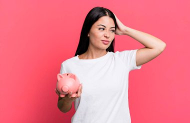 hispanic pretty woman feeling puzzled and confused, scratching head with a piggy bank