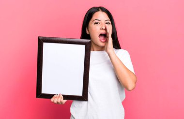 hispanic pretty woman feeling happy,giving a big shout out with hands next to mouth with an empty blank frame