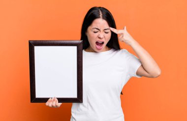 hispanic pretty woman looking unhappy and stressed, suicide gesture making gun sign with an empty blank frame