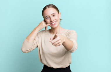 Neşeli bir şekilde gülümsüyor ve telefonla konuşurken kamerayı işaret ediyor.