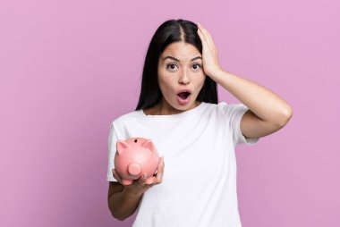 hispanic pretty woman looking happy, astonished and surprised with a piggy bank