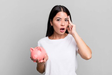 hispanic pretty woman looking surprised, realizing a new thought, idea or concept with a piggy bank