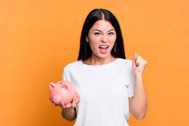 hispanic pretty woman shouting aggressively with an angry expression with a piggy bank