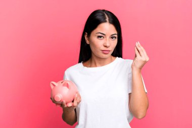 hispanic pretty woman making capice or money gesture, telling you to pay with a piggy bank