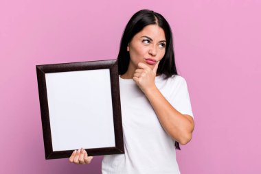hispanic pretty woman thinking, feeling doubtful and confused with an empty blank frame