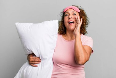 pretty middle age woman feeling happy,giving a big shout out with hands next to mouth wearing pajamas night wear and a pillow
